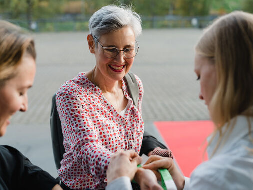 Das Bild zeigt drei Frauen. Eine Frau legt einer anderen ein Armband an.