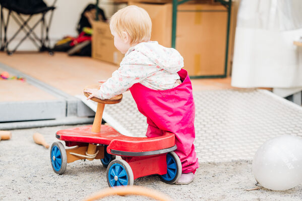 Ein kleines Kind mit roter Latzhose schiebt ein Bobbycar von der Kamera weg.