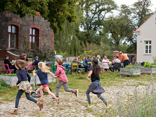 Kinder laufen durchs Bild. Im Hintergrund sitzen Menschen an einem langen Tisch.