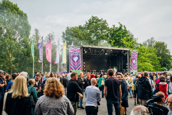 Das Bild zeigt Besucher*innen auf unserem GLS Bank Jubiläumsfestival vor der großen Hauptbühne mit Fahnen im Hintergrund.