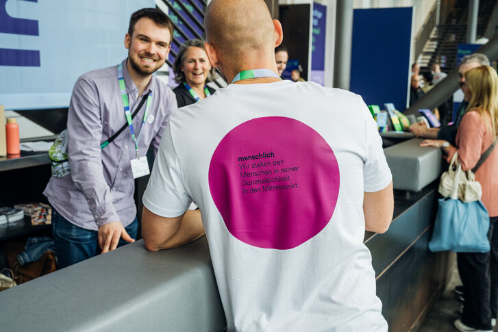 Das Bild zeigt Mitarbeitende im Gespräch. Auf dem Rücken des T-Shirts des Mitarbeiters im Vordergrund steht der GLS Wert "menschlich".