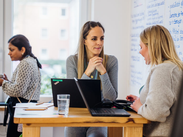Das Bild zeigt zwei Frauen im Austausch. Im Hintergrund ist eine dritte Frau zu sehen.