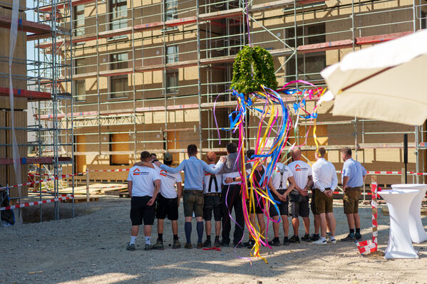Handwerker*innen stehen bei dem Richtfest der Greenfields Buckow vor einem Rohbau in Holzbauweise. Im Vordergrund hängt ein Kranz mit bunten Bändern.