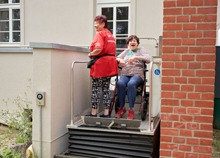 Zwei Frauen nutzen einen Lift, eine Frau sitzt auf einem Rollator.