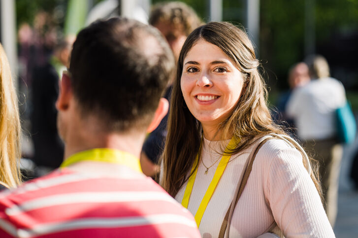 Eine junge Frau schaut in die Kamera. Im Vordergrund sieht man einen Mann von hinten.