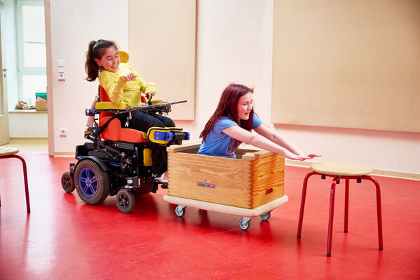 Ein Kind in einem elektrischen Rollstuhl schiebt ein weiteres Kind in einer Rollkiste im Spiel vor sich her. Beide Kinder lachen.