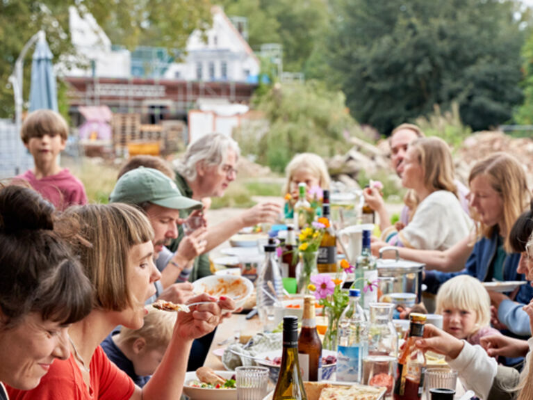 Menschen verschiedenen Alters sitzen an einer langen Tafel zusammen und essen gemeinsam.