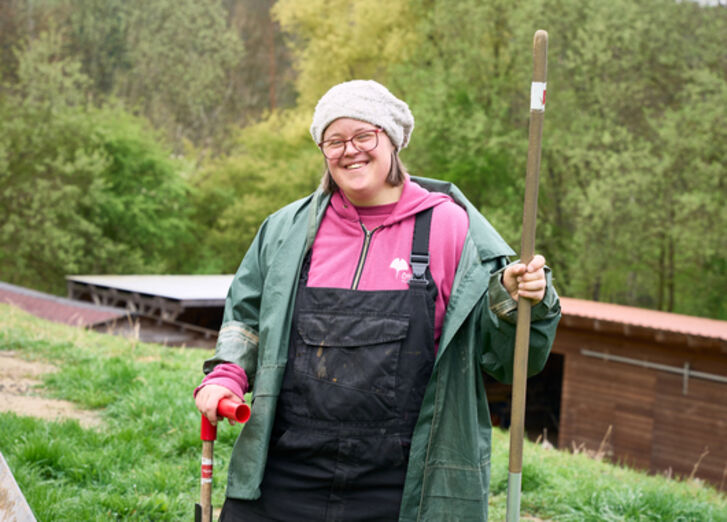 Eine Frau steht lächelnd im Garten und hält ein Gartengerät fest.