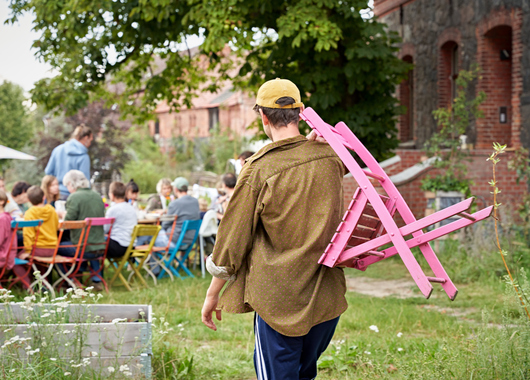 Ein Mann trägt einen Stuhl zu einer langen Tafel im Garten, an der bereits Menschen verschiedenen Alters zusammen sitzen.